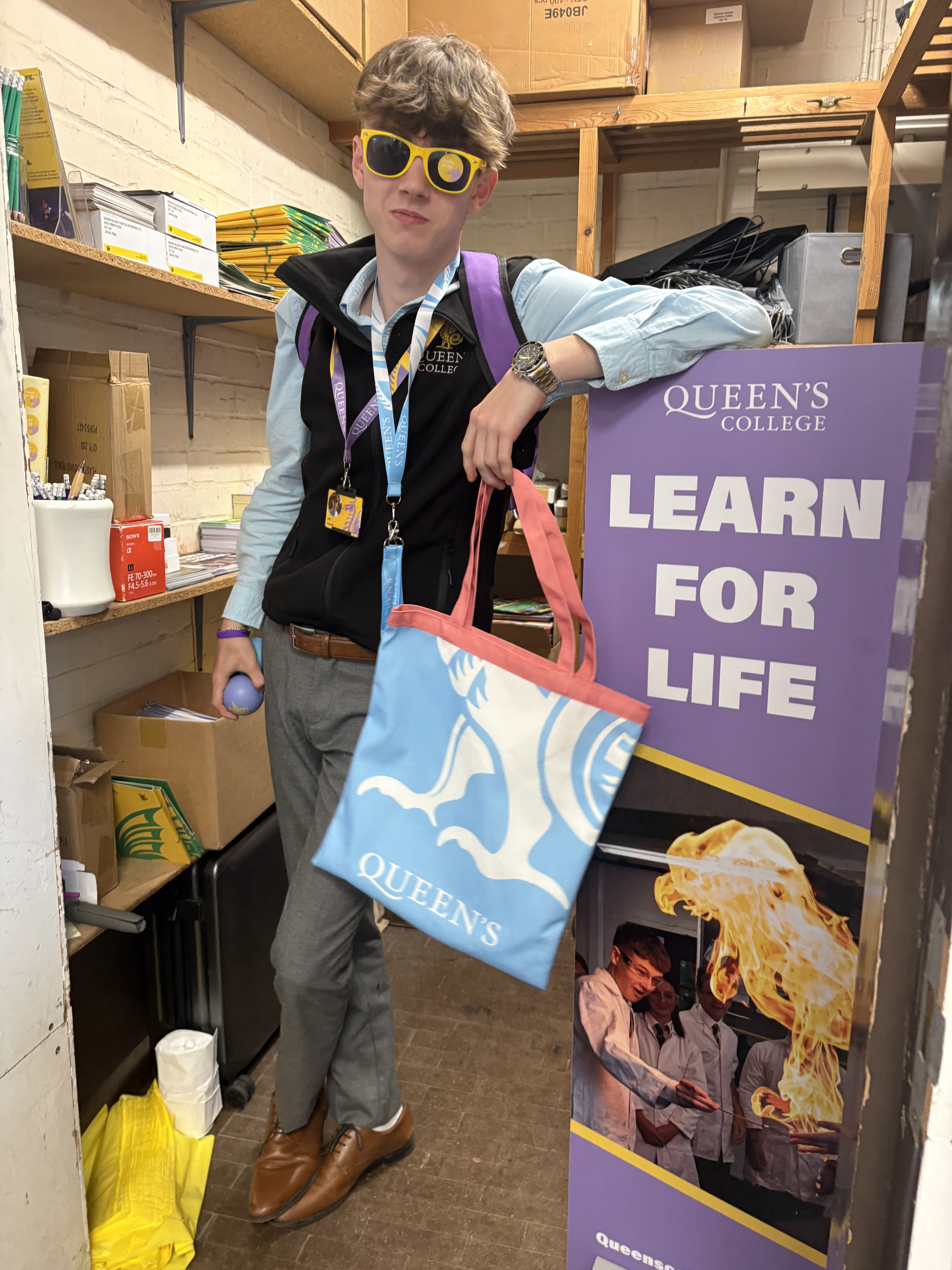 Jack Frampton modelling Queen's College branded merchandise — tote bag, lanyard, sunglasses and stress ball — beside a Learn for Life pull-up banner